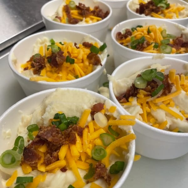 Multiple white cups filled with mashed potatoes, topped with shredded cheese, bacon bits, and green onions.